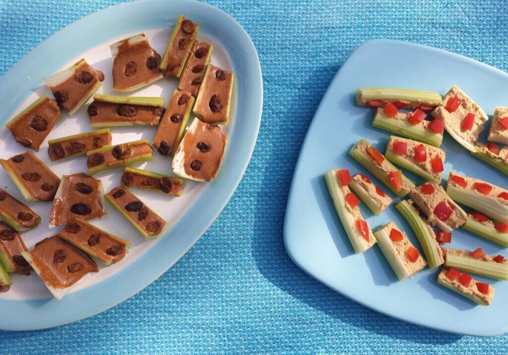 bleekselderijstengels gevuld met pindakaas en rozijnen en met hummus en stukjes rode paprika